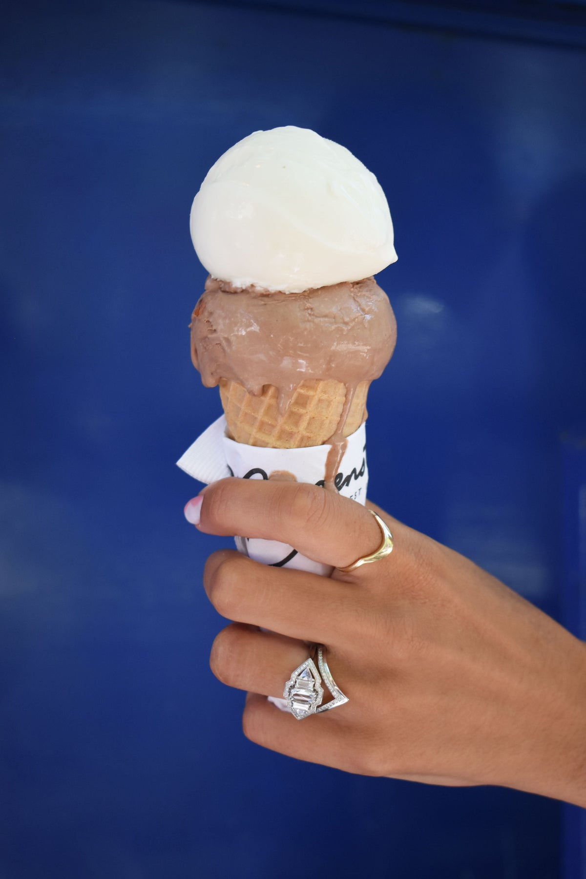 A photo of a woman's hand holding a Morgenstern's ice cream cone with chocolate and vanilla ice cream scoops against a blue background. She is wearing Anna Sheffield fine jewelry rings in white gold and yellow gold on her index and ring fingers. 