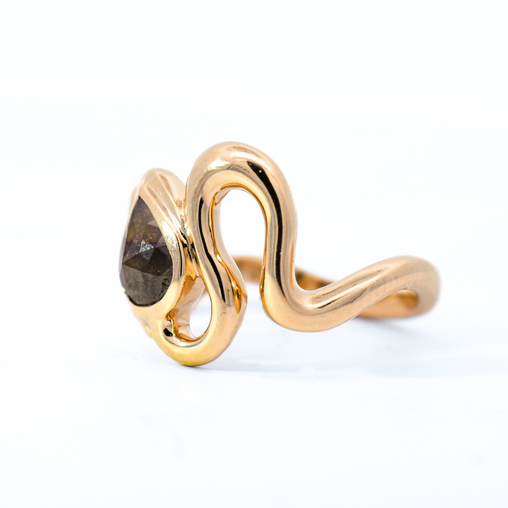 Side profile of a ring inspired by the organic curving silhouette of a serpent featuring a brown pear cut diamond set in yellow gold against a white background. 