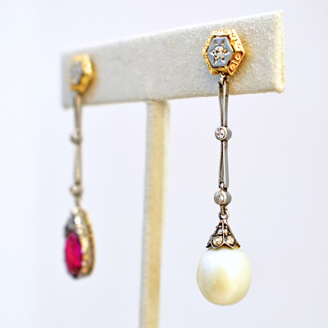 Two drop earrings on a white suede stand. A hexagon shaped white gold post featuring a star engraving and encircled in yellow gold drop down into white gold chain links separated by white round diamonds to a round pearl droplet. The earring on the left is the same design except it features a ruby droplet instead of a pearl. 