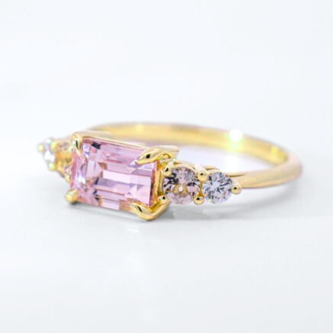 A 3/4 angle of a five stone ring featuring an emerald cut pink morganite center stone and one round morganite and one round white diamond on each side set in yellow gold against a white background. 