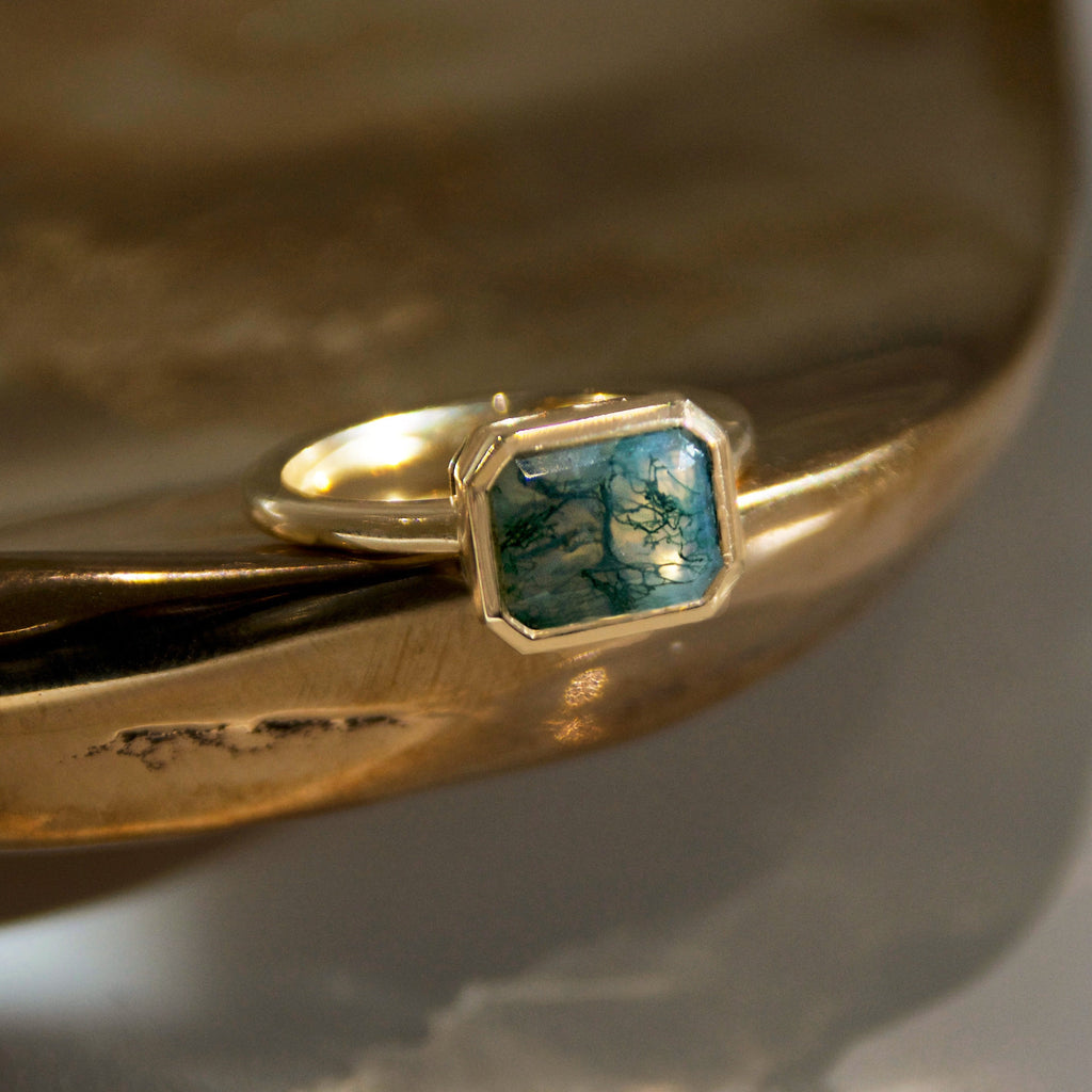 A yellow gold ring featuring a moss agate emerald cut stone set in a gold bezel on a golden bronze dish 