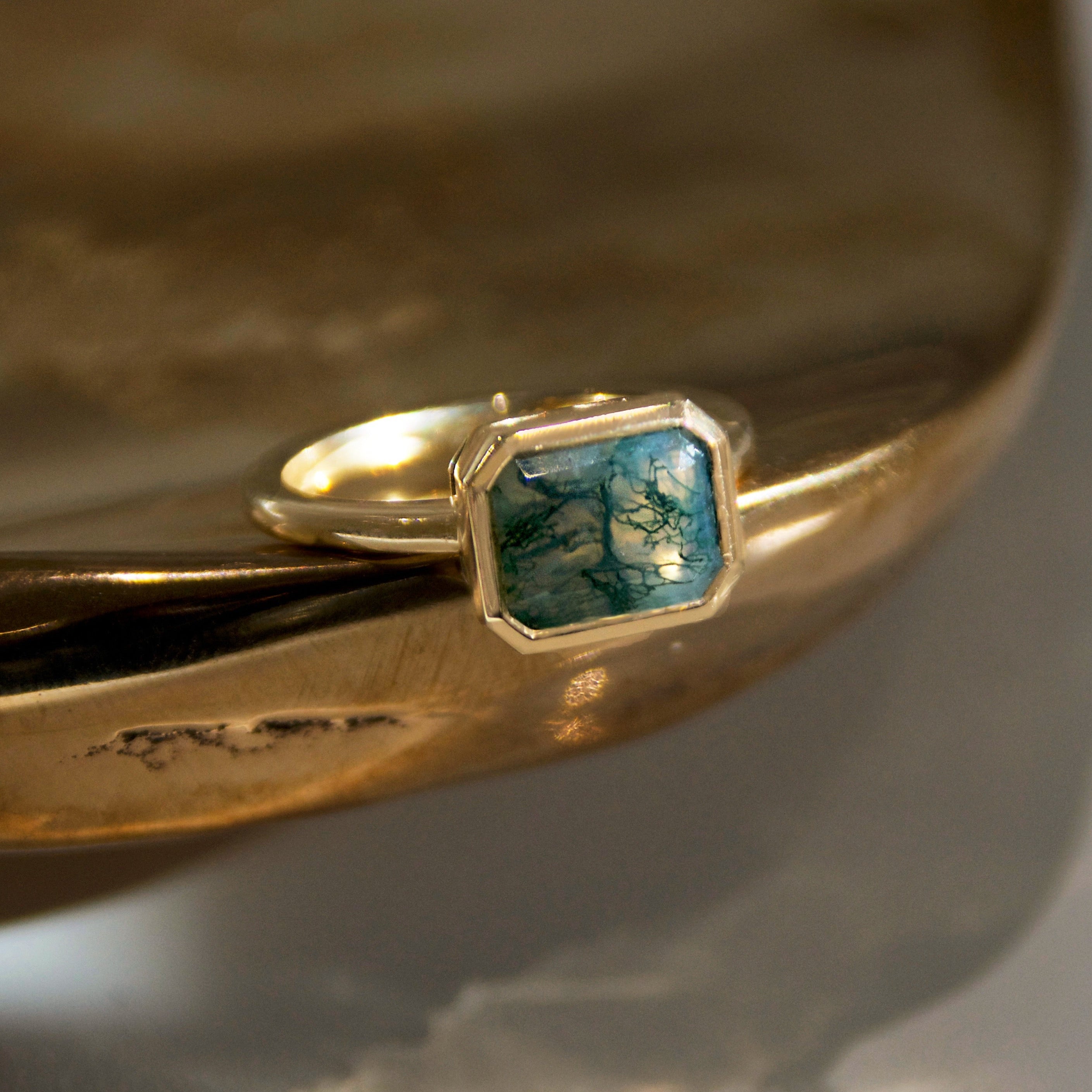A yellow gold ring featuring a moss agate emerald cut stone set in a gold bezel on a golden bronze dish 
