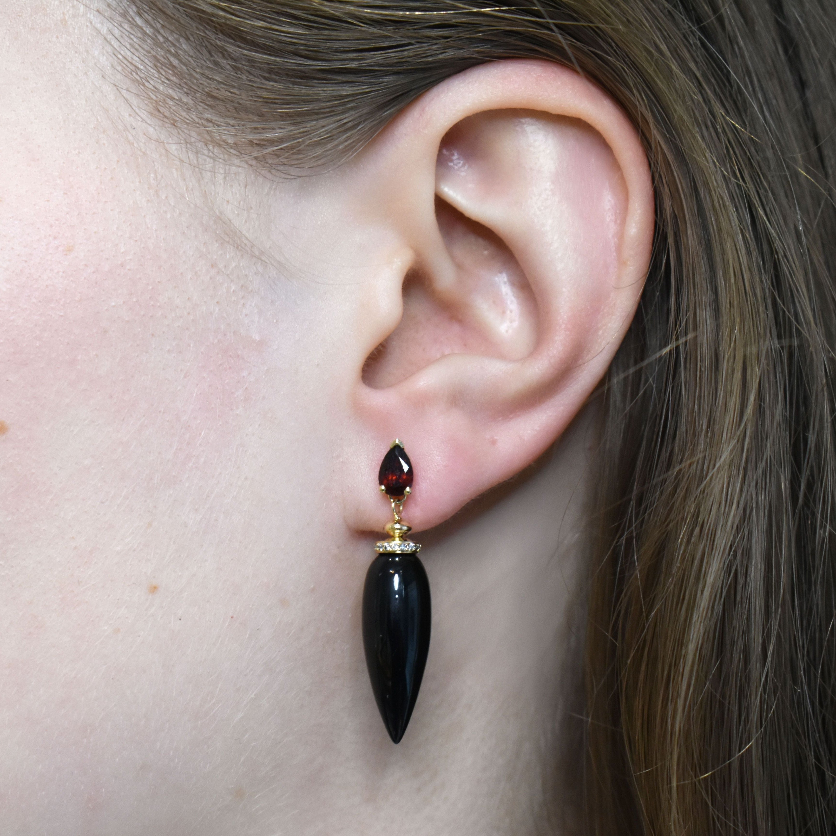 A black onyx pendulum earring featuring white diamonds and red garnet set in yellow gold on an ear. 
