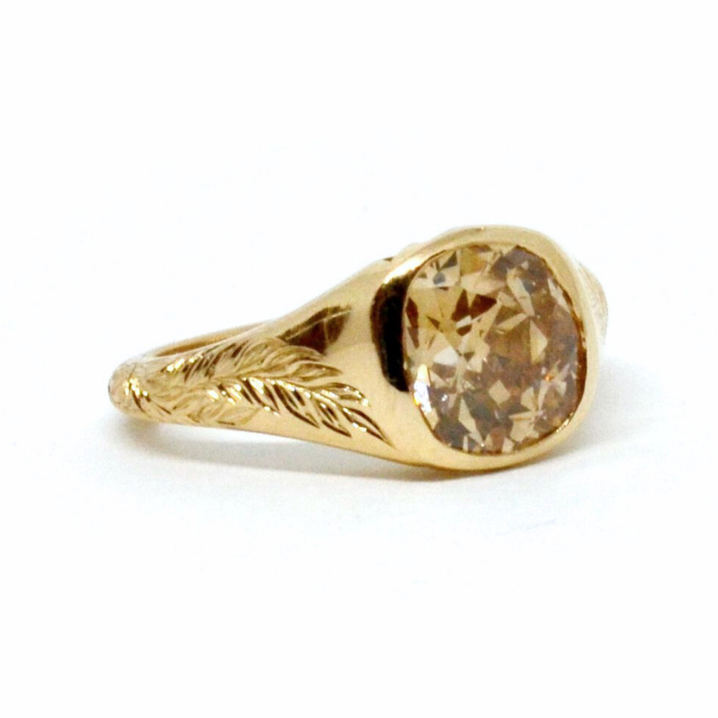 A 3/4 angle of a yellow gold signet ring featuring a cushion cut brown diamond center and leaf engravings on both sides of the band against a white background.