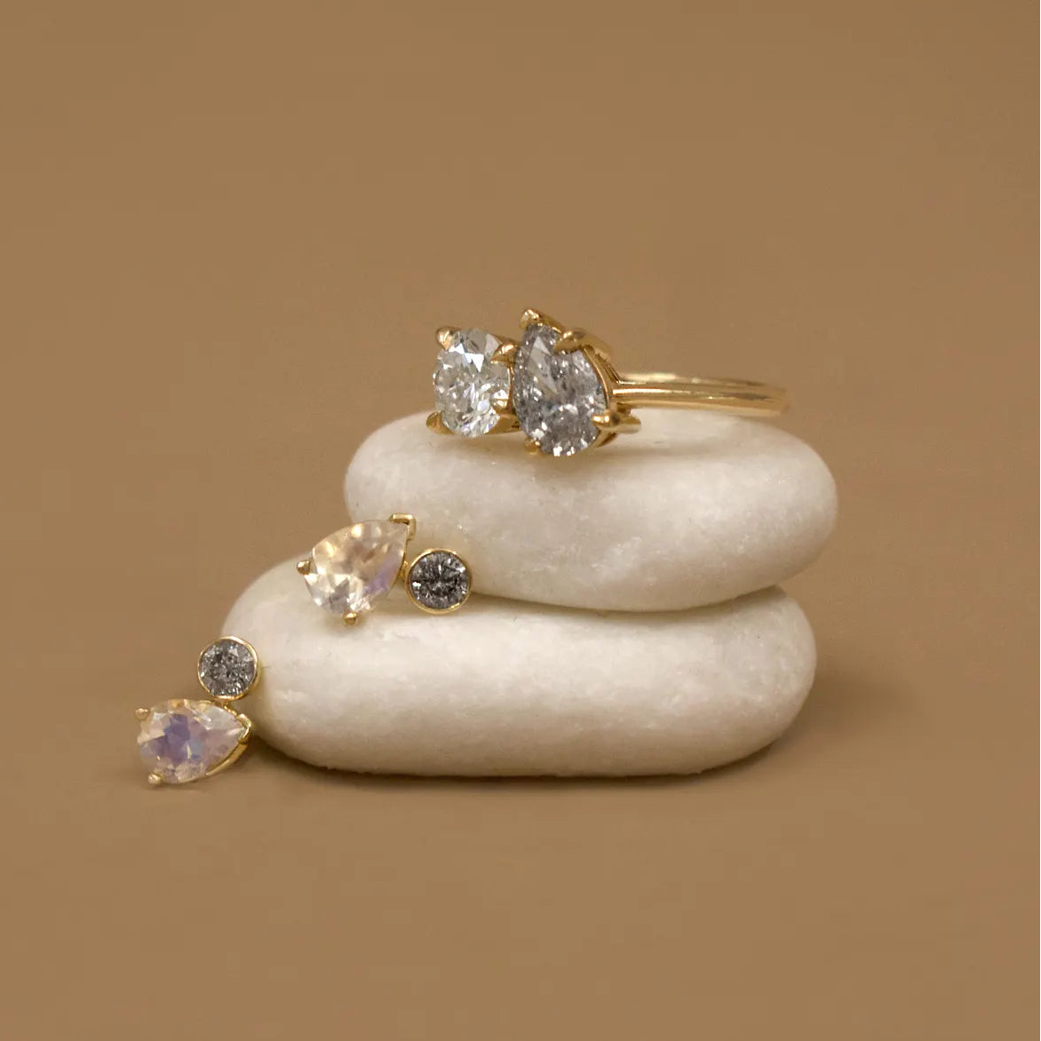 Two moonstone and diamond stud earrings and a toi et moi white and grey diamond gold ring perched on two white stacked stones on a tan brown background 