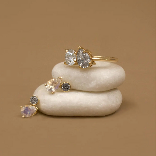 Two moonstone and diamond stud earrings and a toi et moi white and grey diamond gold ring perched on two white stacked stones on a tan brown background 