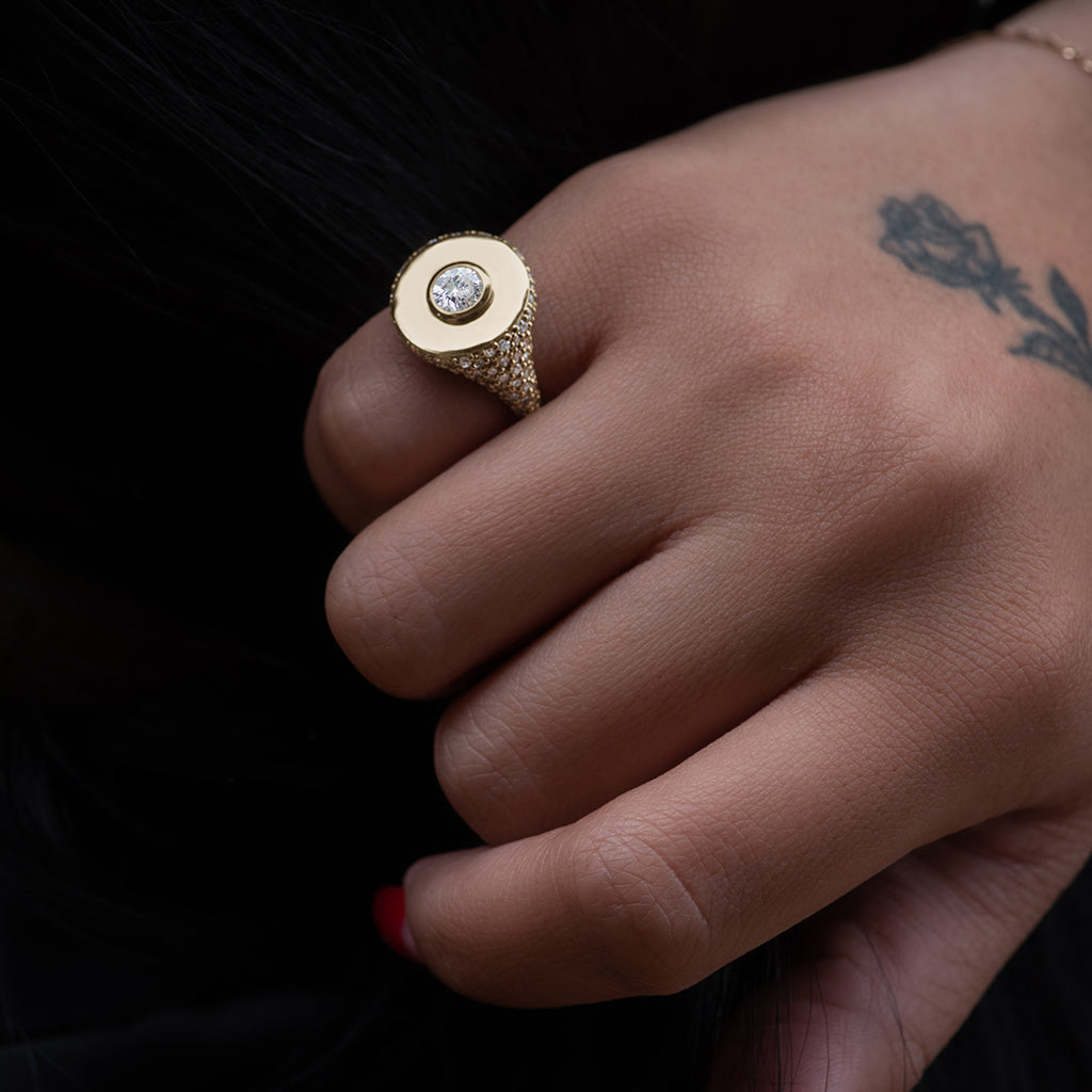 A round white diamond bezel set in a signet style ring with white diamond pave down the sides of the band set in yellow gold on a medium-complexion model's pinky finger.