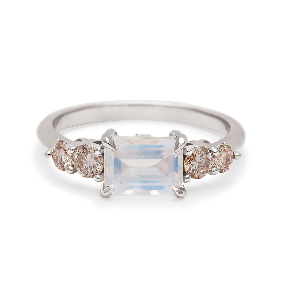 A ring with an emerald cut and shaped rainbow moonstone center stone with two round champagne diamonds on either side set in white gold., White Gold