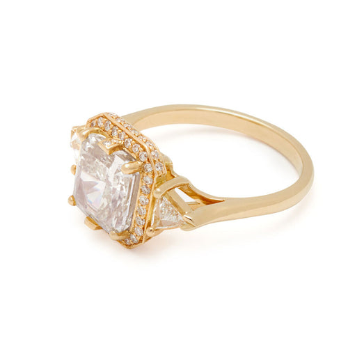 A radiant cut lab white diamond encircled by a white diamond halo and two white lab trillion diamond side stones set in a yellow gold ring on a white background 