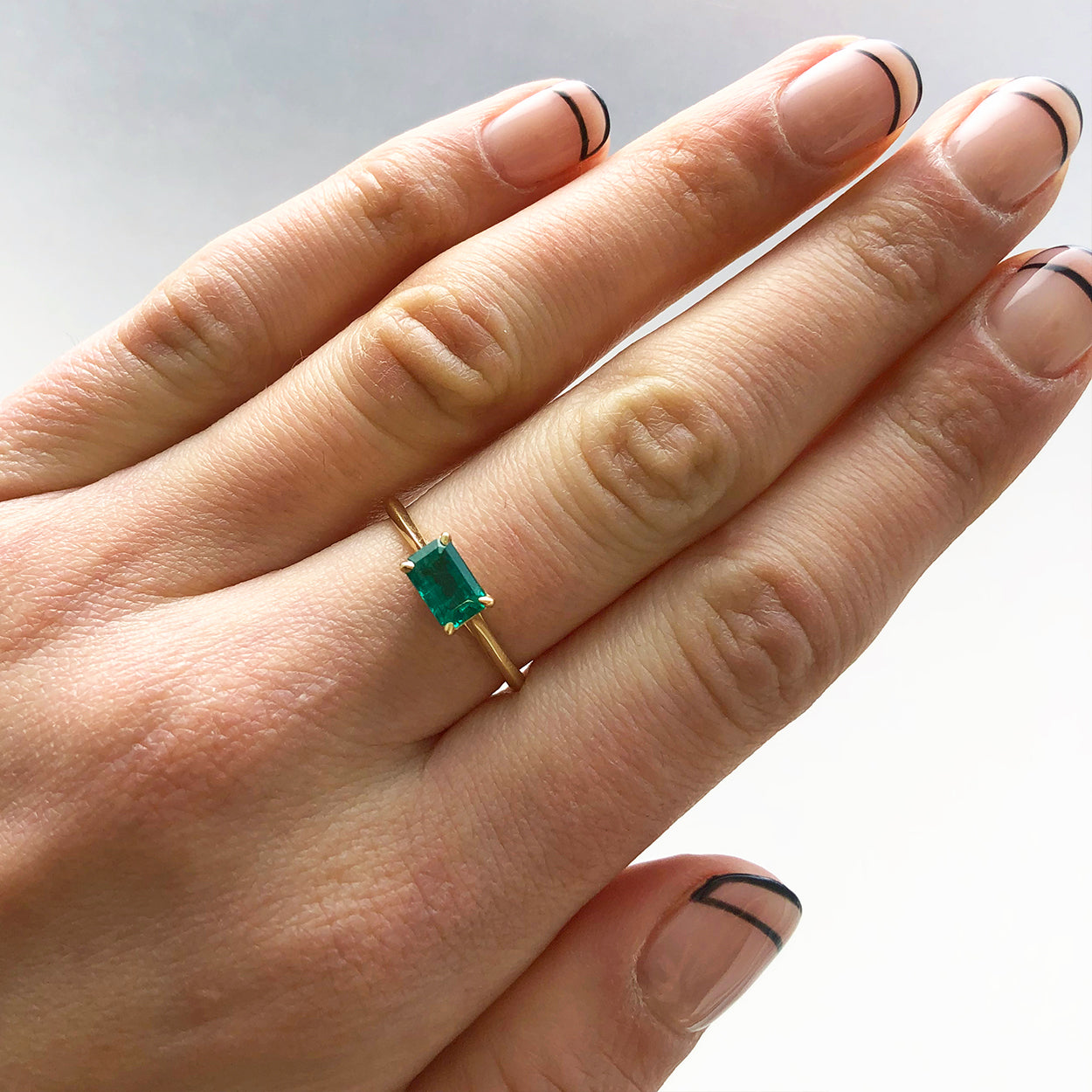 An east/west solitaire ring with an emerald shape green emerald stone set in yellow gold. On a finger with linear polish., White Gold, Rose Gold, Yellow Gold