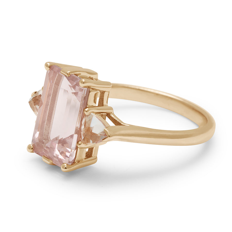 Angled view of a three stone engagement ring with an emerald shape pink morganite center stone with two peach morganite trillion side stones set in yellow gold.