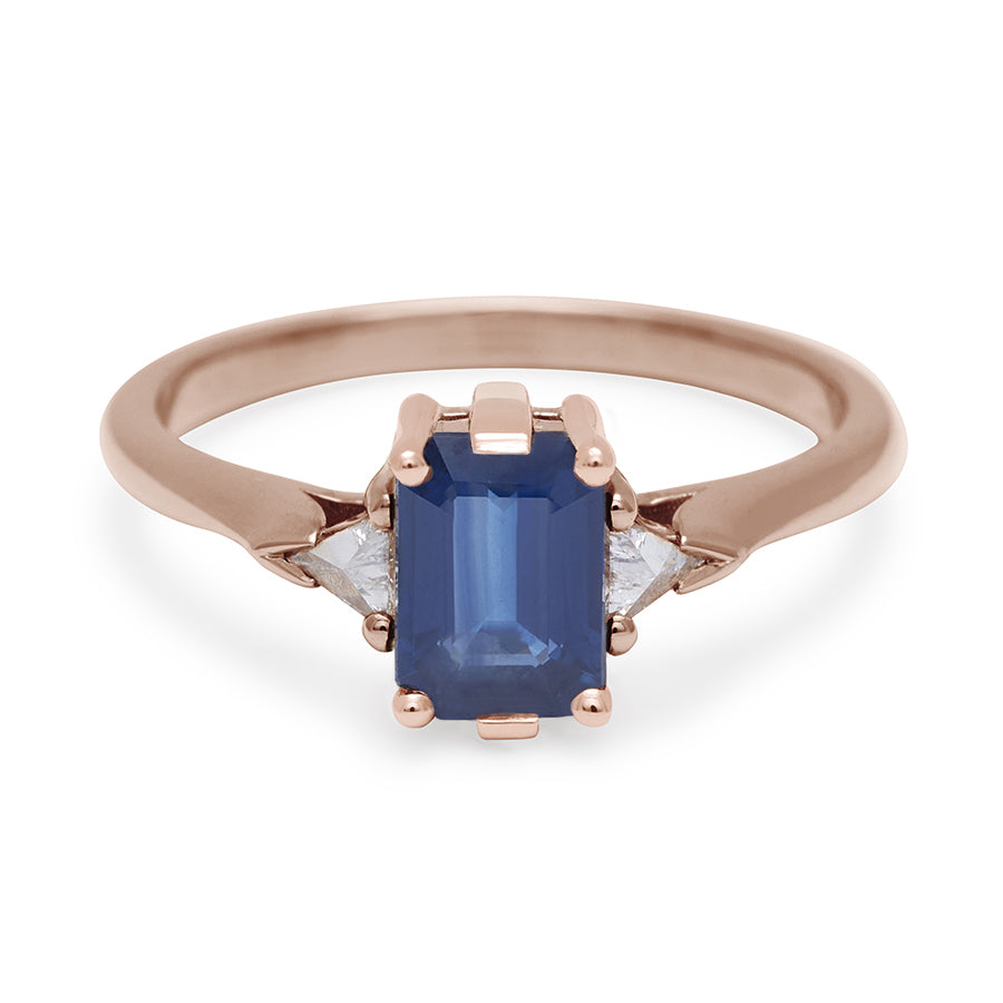A three stone engagement ring with an emerald cut and emerald shape 7x5mm blue sapphire center stone with two white diamond trillion side stones set in rose gold., Rose Gold