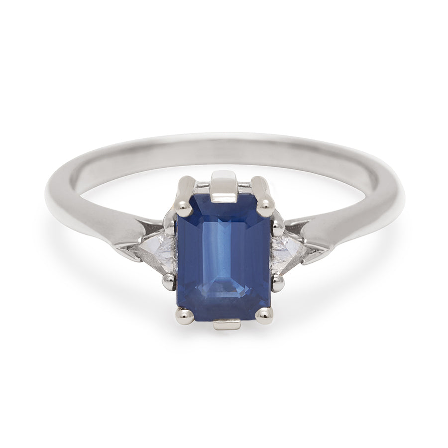 A three stone engagement ring with an emerald cut and emerald shape 7x5mm blue sapphire center stone with two white diamond trillion side stones set in white gold., White Gold