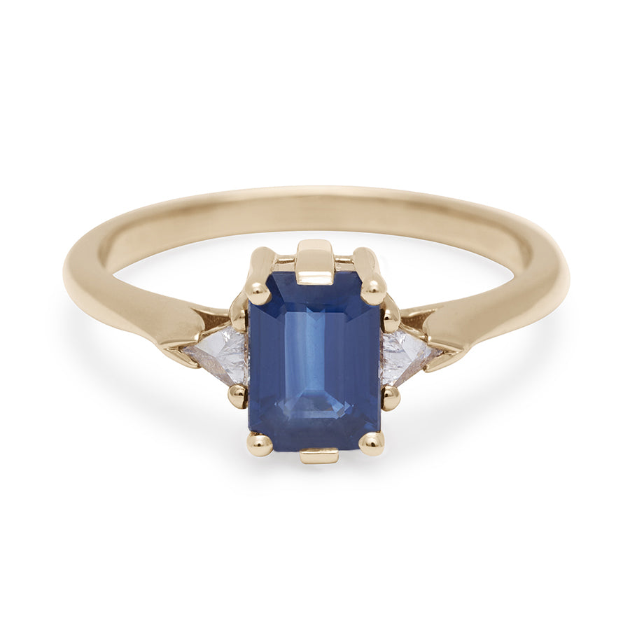 A three stone engagement ring with an emerald cut and emerald shape 7x5mm blue sapphire center stone with two white diamond trillion side stones set in yellow gold., Yellow Gold