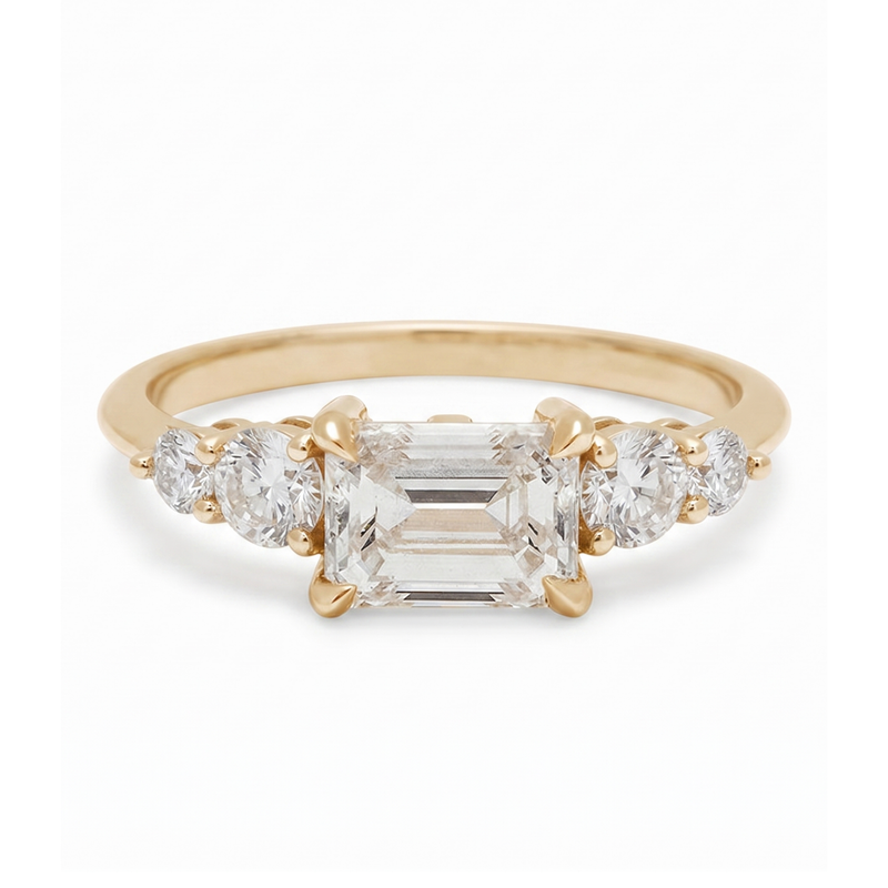 Gold ring with a central emerald-cut diamond and two side stones on a white background