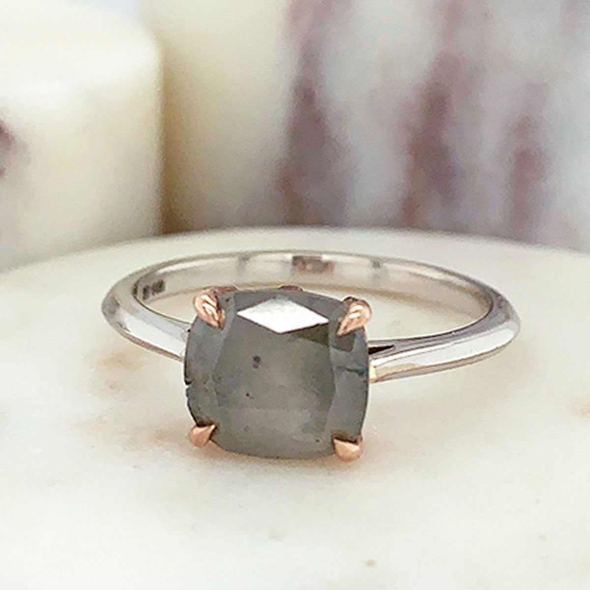 An east/west solitaire ring with an oval shape grey diamond stone set in a rose gold basket and a white gold band on a marble stone.