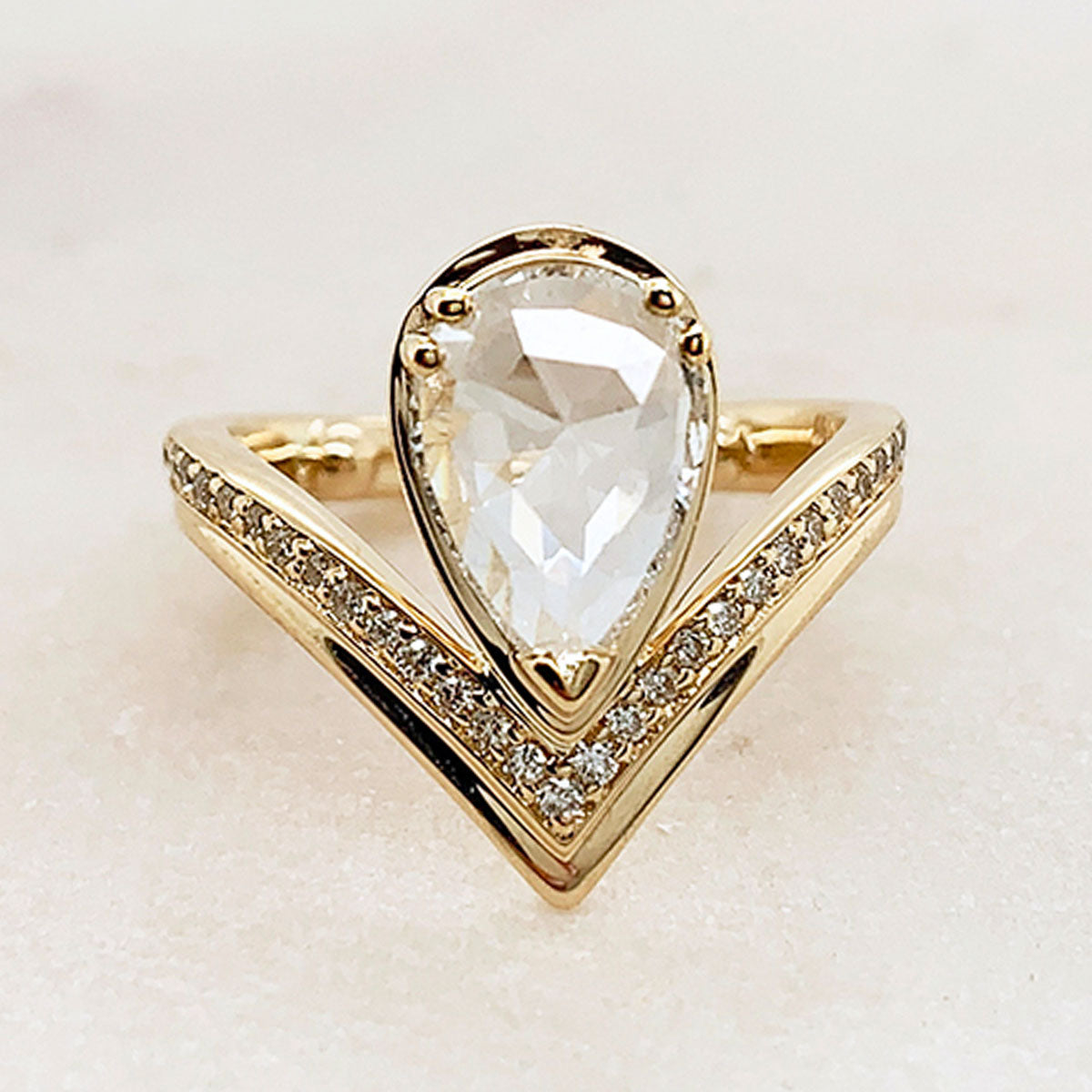 A pear shaped rainbow moonstone set above a white diamond dusted band set in yellow gold on a grey background.
