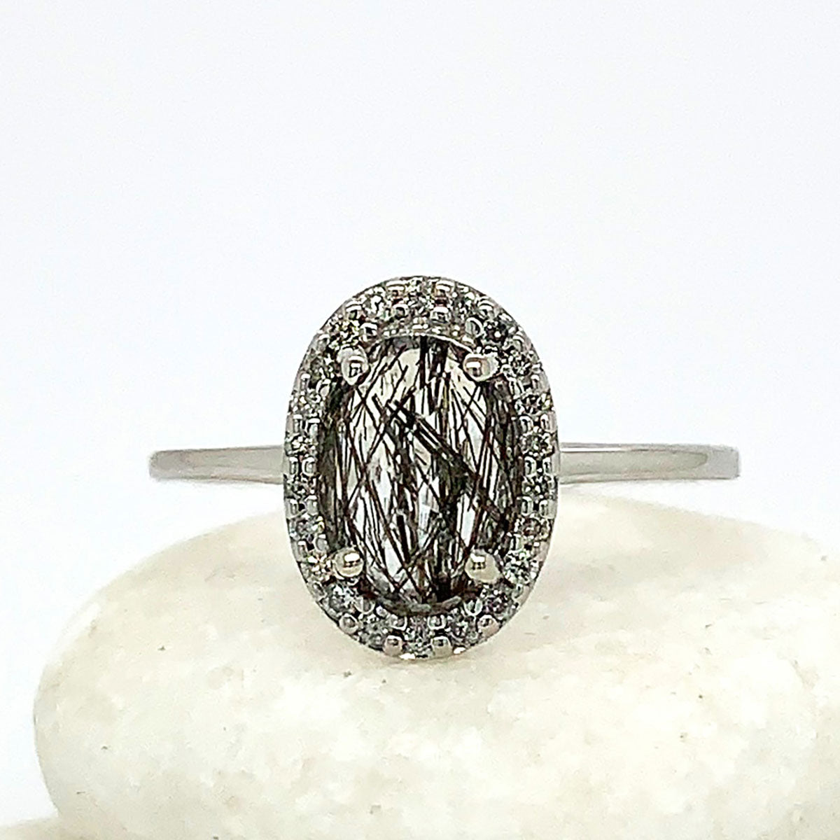 An engagement ring with an oval shape black rutilated quartz center stone surrounded by a white diamond pave halo set in white gold band on three white stones.