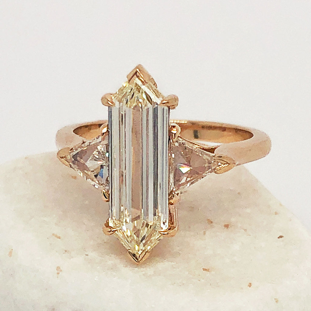 A three stone engagement ring with an hexagonal shape white diamond center stone with two white diamond trillion side stones set in yellow gold on a white rock.
