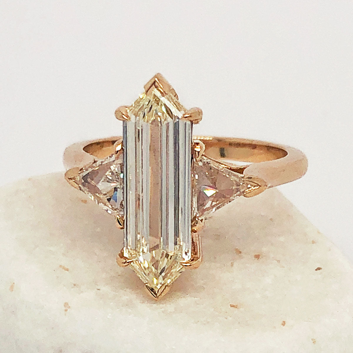 A three stone engagement ring with an hexagonal shape white diamond center stone with two white diamond trillion side stones set in yellow gold on a white rock.