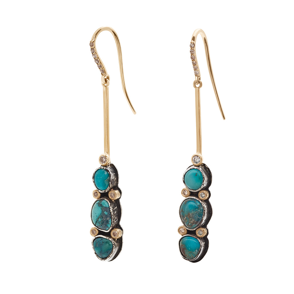 Angled view of a pair of drop earrings with turquoise stones set in sterling silver and champagne diamond pave accent stones and on the earwire set in yellow gold.