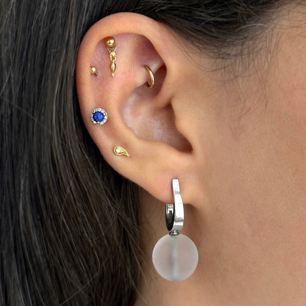 Close-up of a person's ear showcasing multiple ear piercings and jewelry, including several gold cartilage studs, a blue sapphire stud, a gold hoop, and a silver hoop with a round milky white disc drop earring.