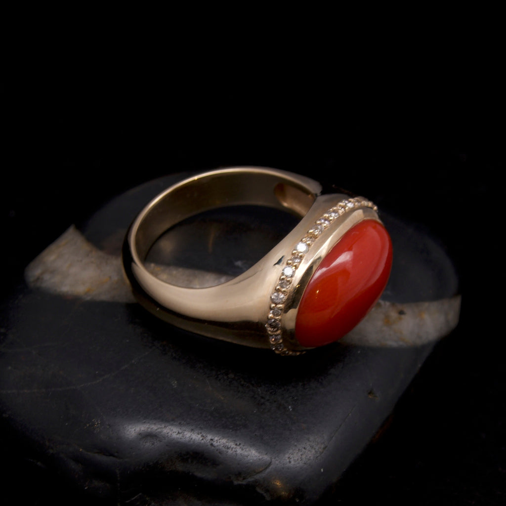 A gold ring with a round red cabochon coral and small clear stones in a bezel setting, displayed on a dark stone against a black background.