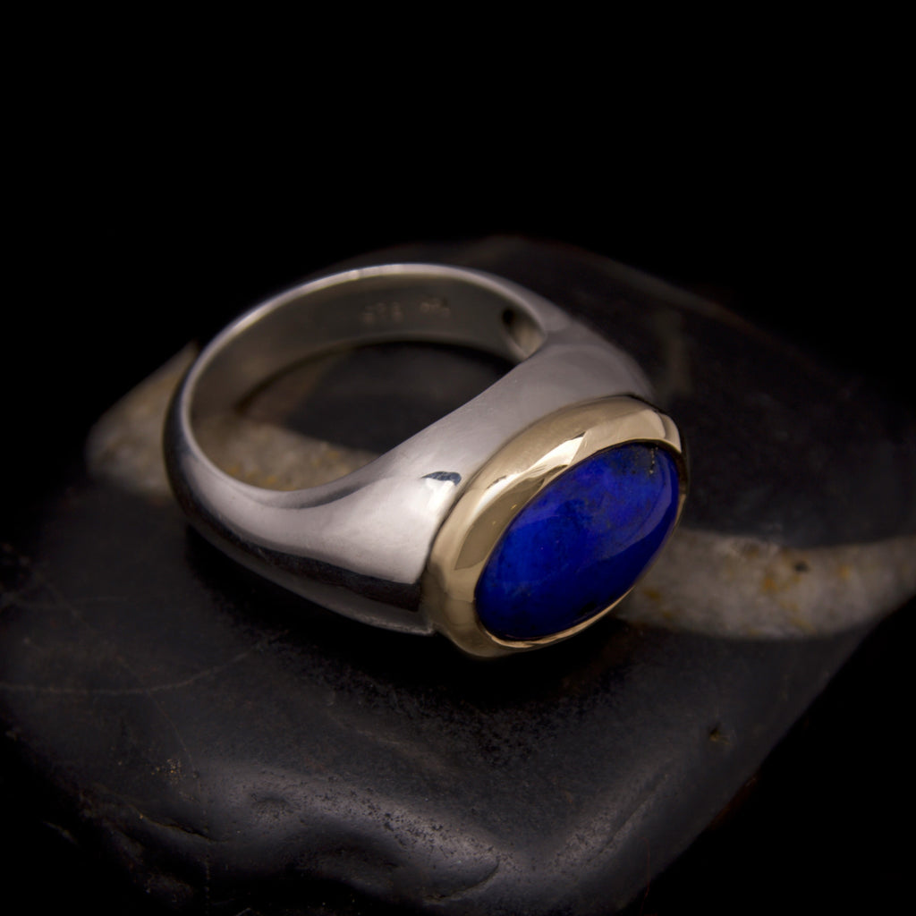 A silver ring with a gold-set deep blue oval gemstone, displayed on a dark polished stone against a black background.