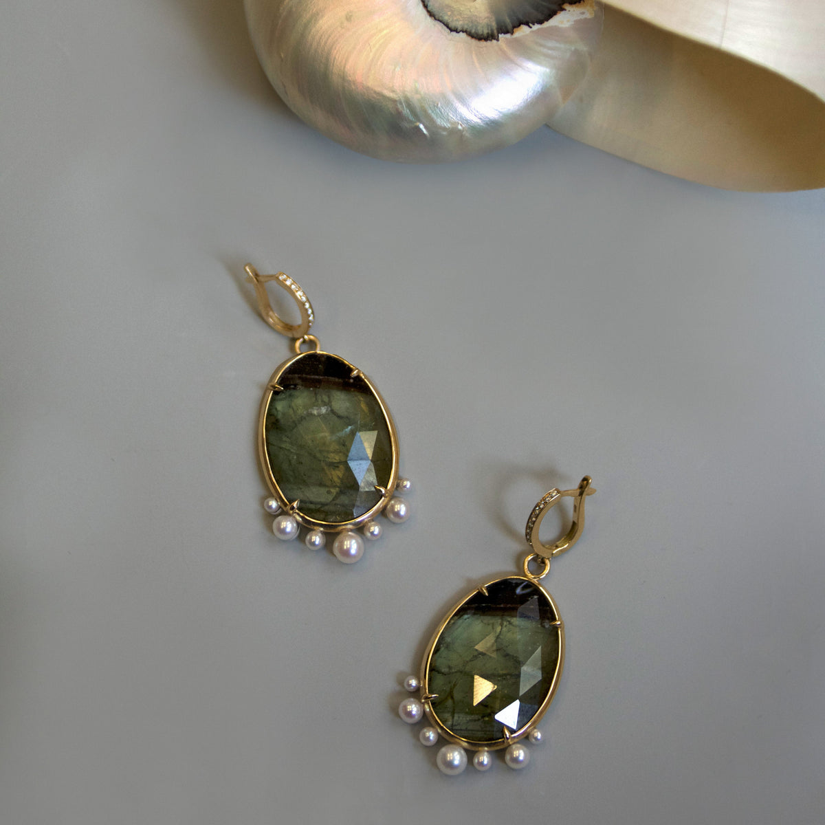 A pair of gold drop earrings with faceted olive green gemstones and small white pearls, displayed on a neutral gray surface next to a large white seashell.