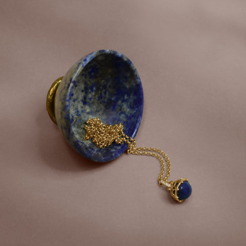 Blue lapis stone bowl with a gold chain with a lapis charm on a beige background