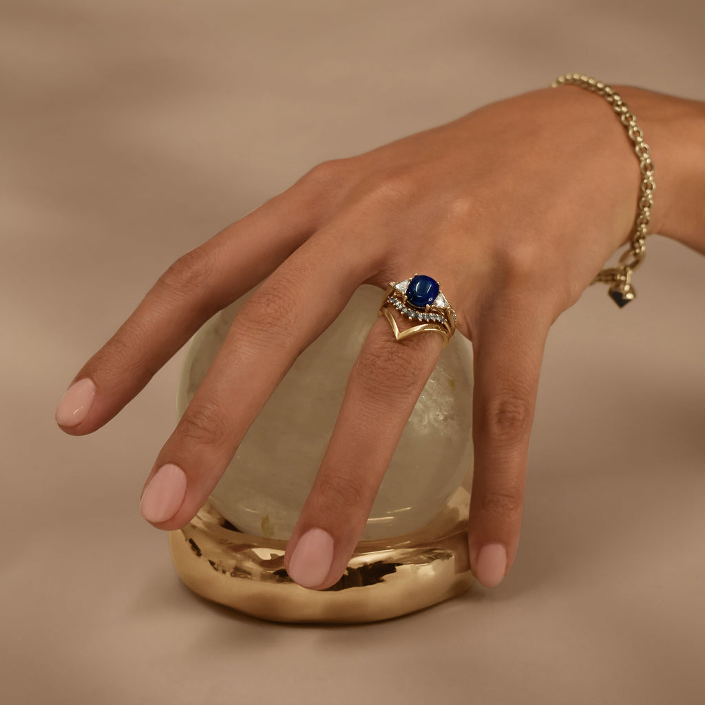 A hand wearing a gold bracelet and several rings on a crystal ball in a gold stand against a beige background 