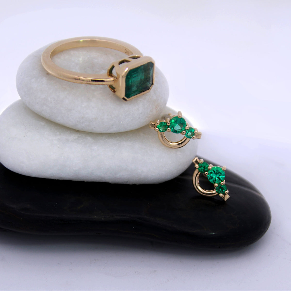 A stack of three white stones and one black stone on a white surface, displaying a gold ring with a green rectangular gemstone and matching gold earrings with green stones.