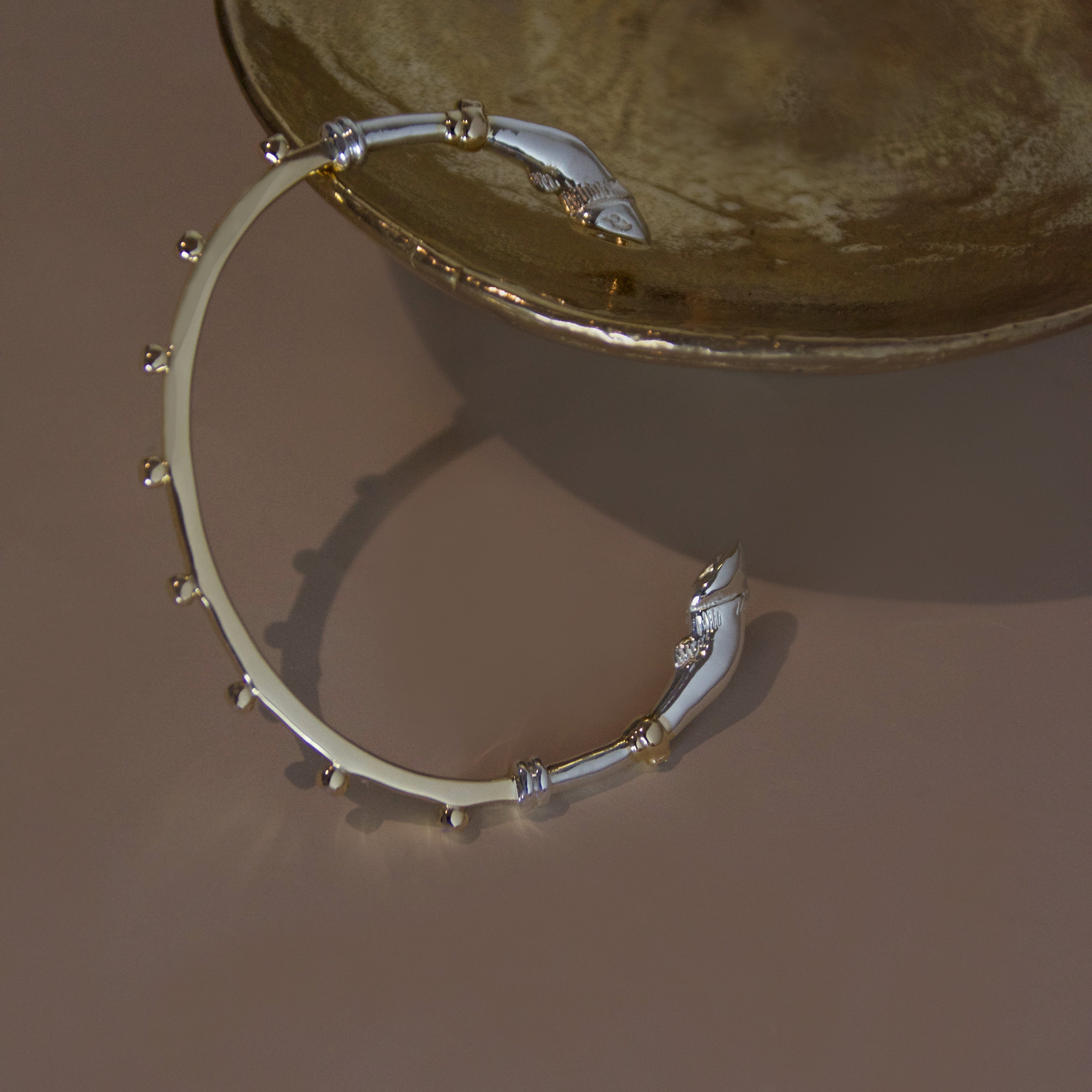 A mixed metal yellow gold and sterling silver bracelet cuff with horse hoof details against a bronze object on a beige background 