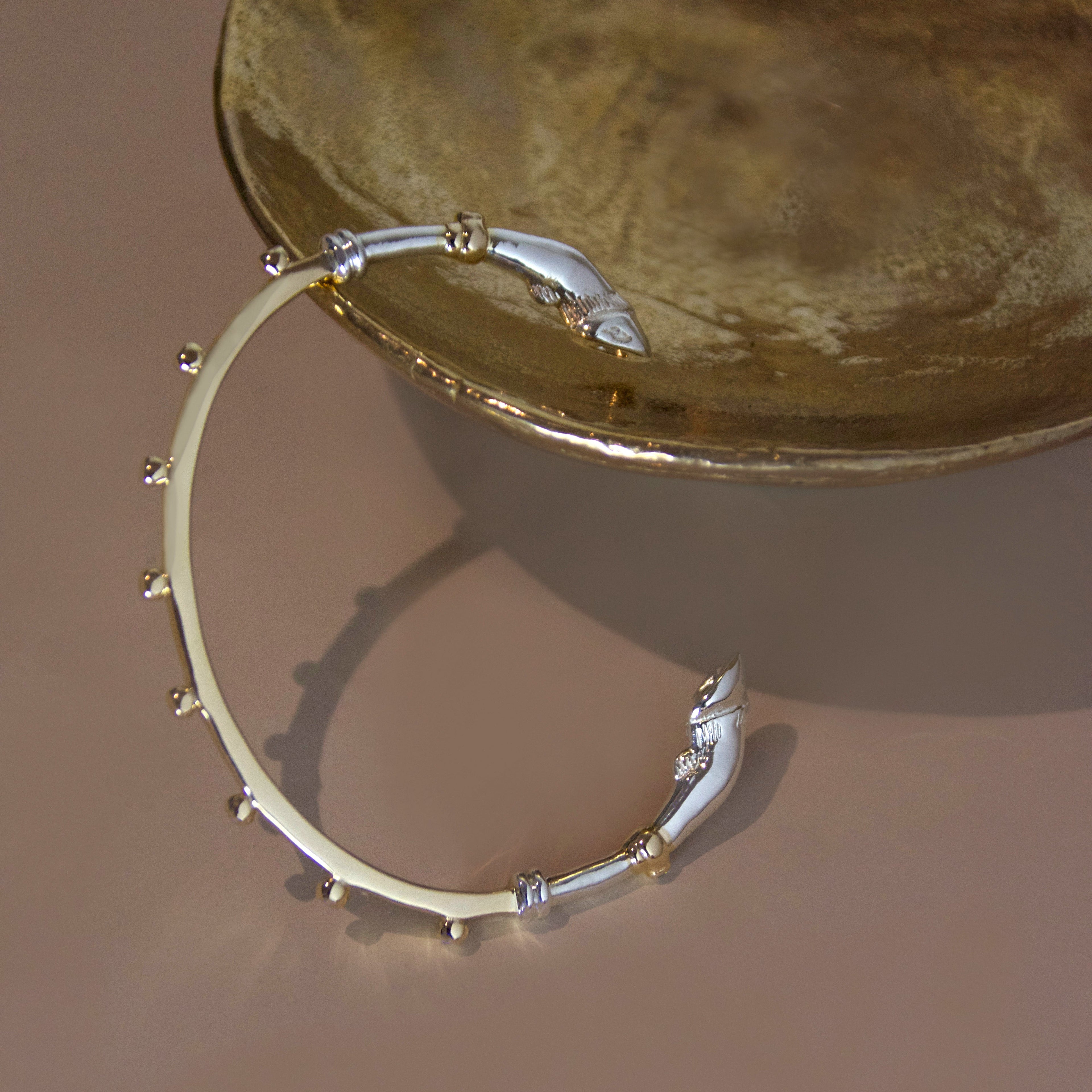 A mixed metal yellow gold and sterling silver bracelet cuff with horse hoof details against a bronze object on a beige background 