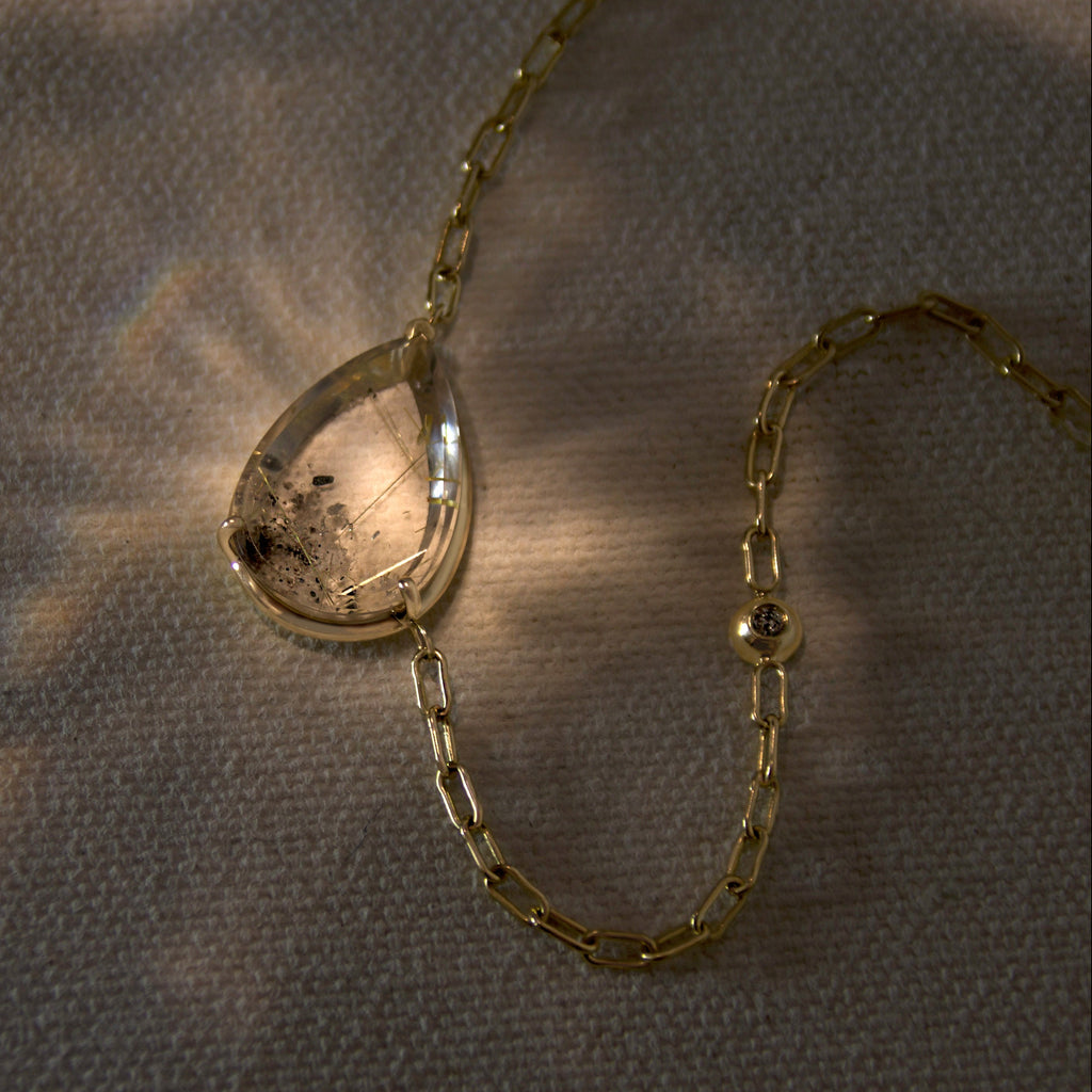 A pear shaped quartz necklace set in yellow gold on a beige canvas background with light shining on it 