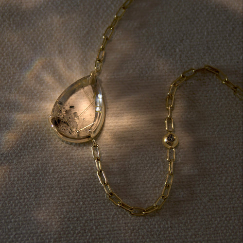 A pear shaped quartz necklace set in yellow gold on a beige canvas background with light shining on it 