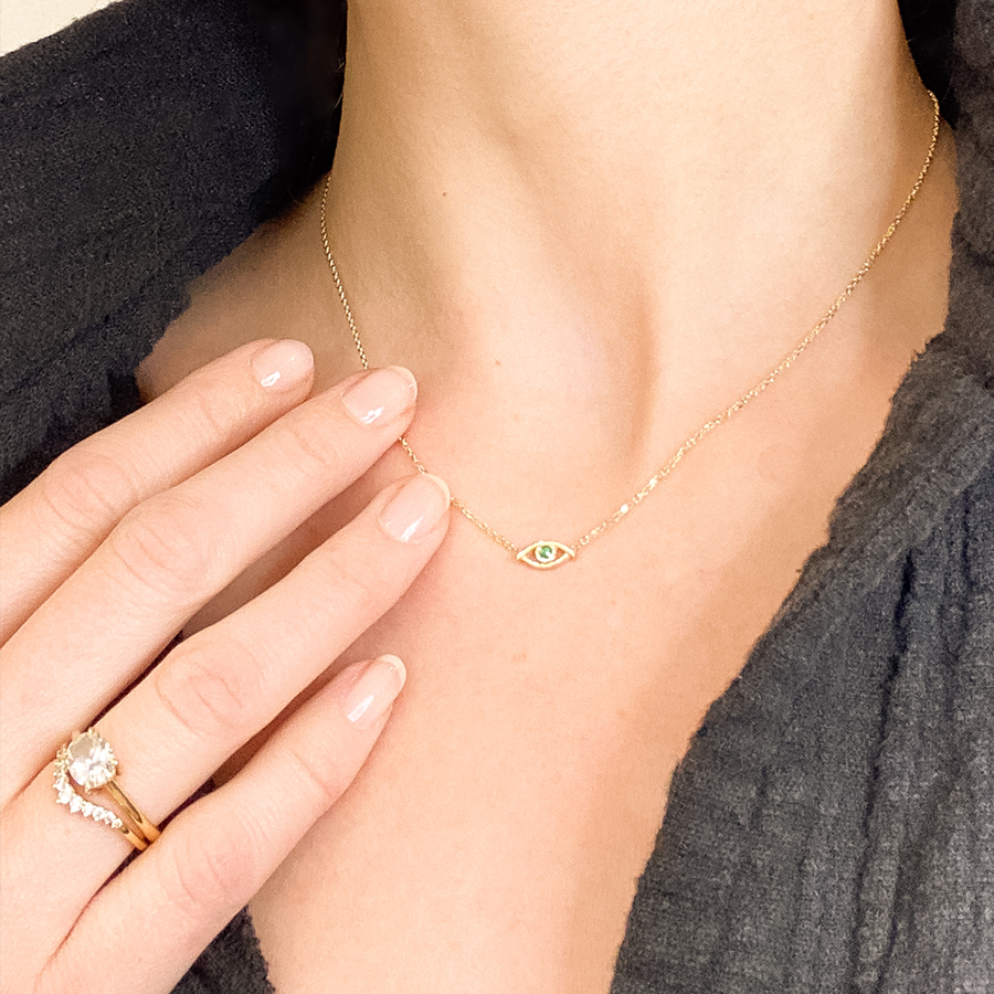 A choker with an evil eye charm with an emerald stone in the center set on a yellow gold chain on a light-complexion model's neck.