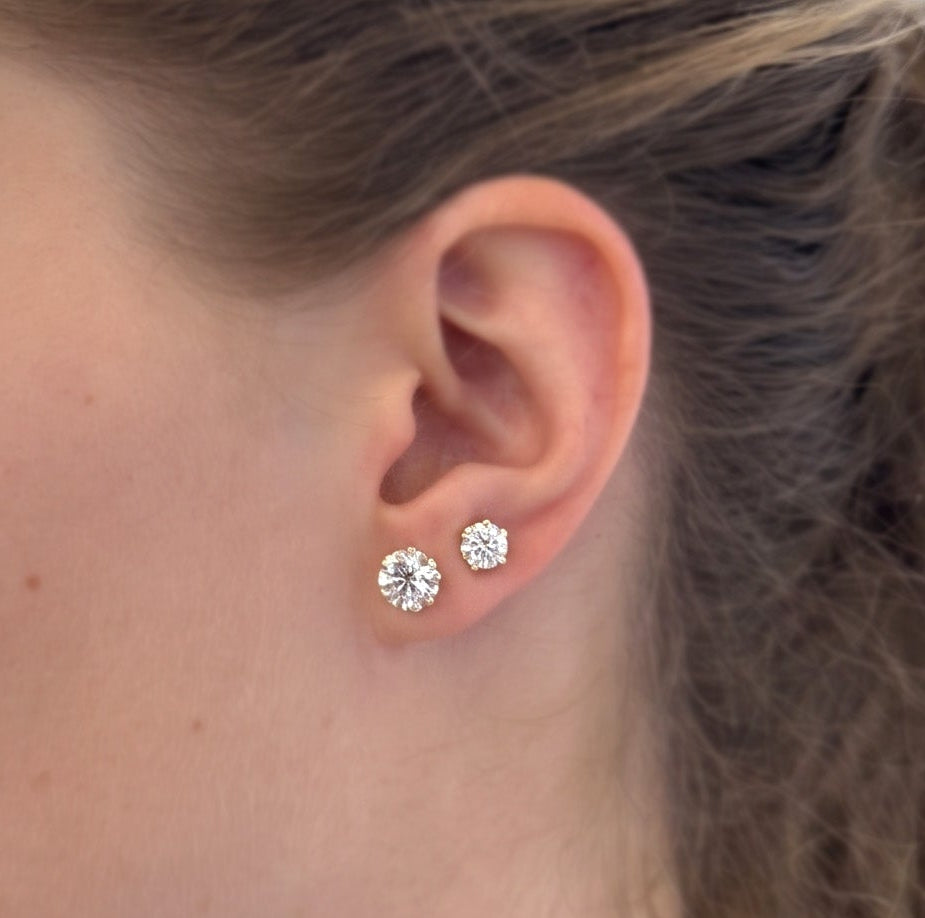 A woman in profile wearing a pair of small round-cut silver stud earrings with clear stones, shown in a brightly lit indoor setting.
