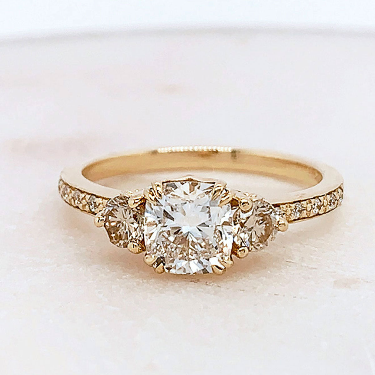 A three stone engagement ring with a cushion cut white diamond center stone with two champagne diamond round side stones set in yellow gold band with white diamond pave around it on a grey background.