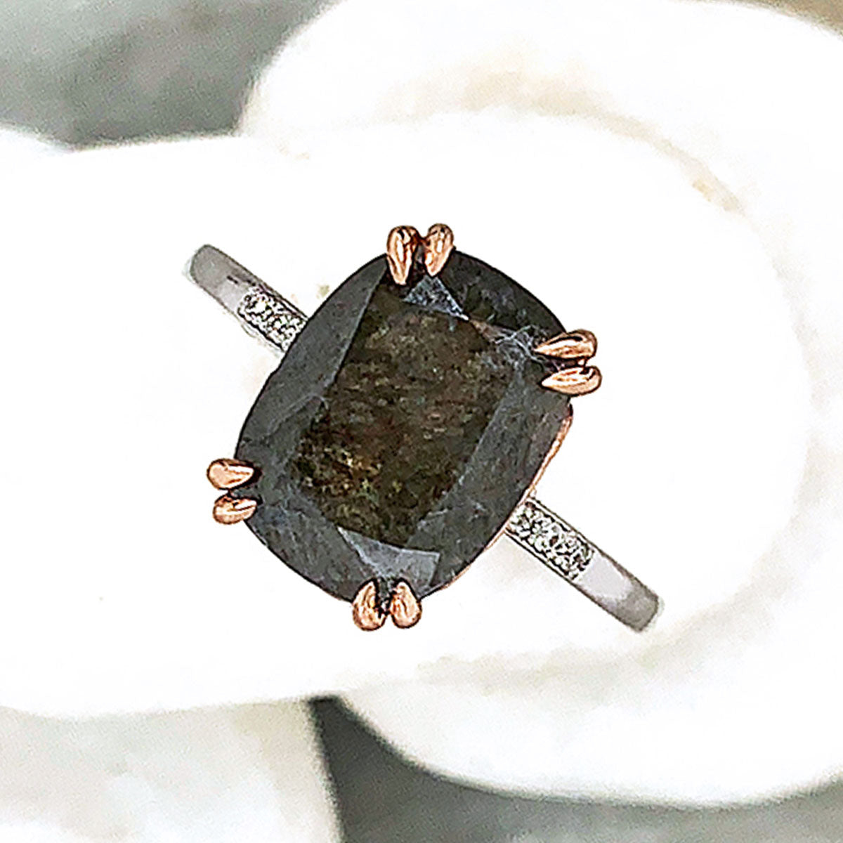 A solitaire ring with a cushion cut grey diamond with white diamond pave on either side set in rose gold basket and white gold band on three white stones.