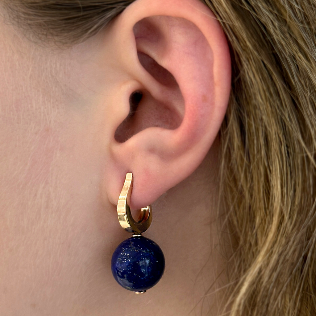 A woman's ear with a yellow gold hook hoop earring with a blue lapis gemstone 