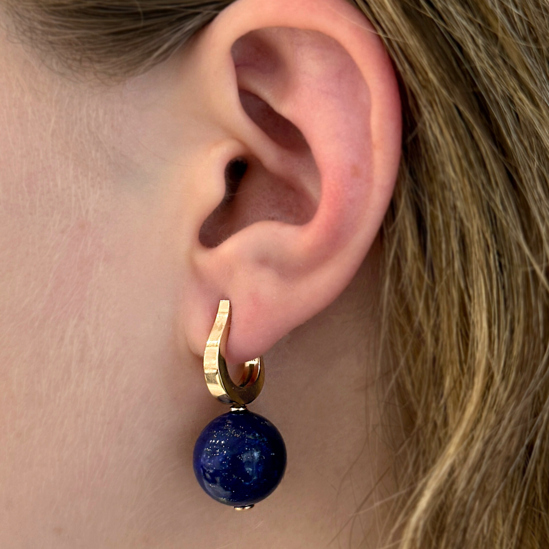 A woman's ear with a yellow gold hook hoop earring with a blue lapis gemstone 