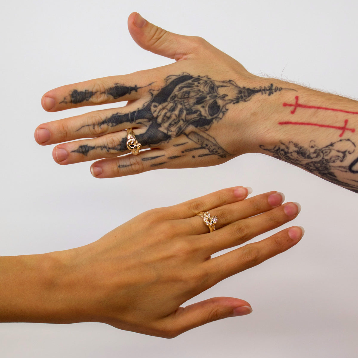 Two hands shown against a plain light background, each wearing a gold ring. The upper hand has a large black tattoo with a skull and crossbones design and red cross markings on the wrist, while the lower hand is clean and un tattooed.