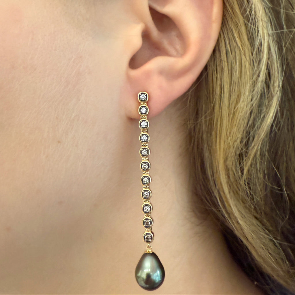 A woman's ear wearing a yellow gold drop earring featuring white and champagne diamonds bezeled followed by a bluish-greenish dark Tahitian pearl droplet