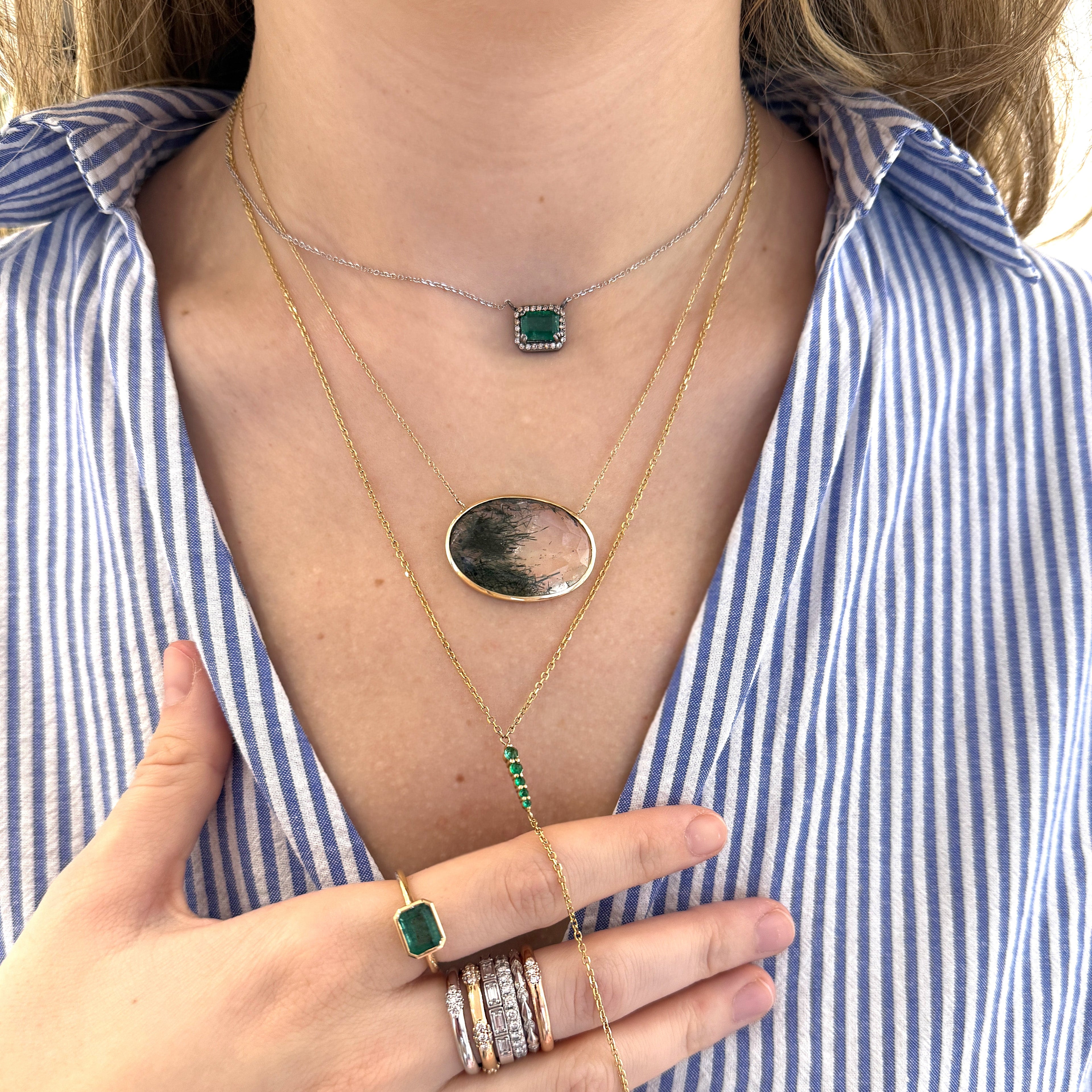A woman wearing three different necklaces, starting with one smaller emerald gemstone necklace with white diamonds, followed by a larger necklace with an oval quartz pedant, and lastly a delicate long green emerald y-shaped gold necklace.