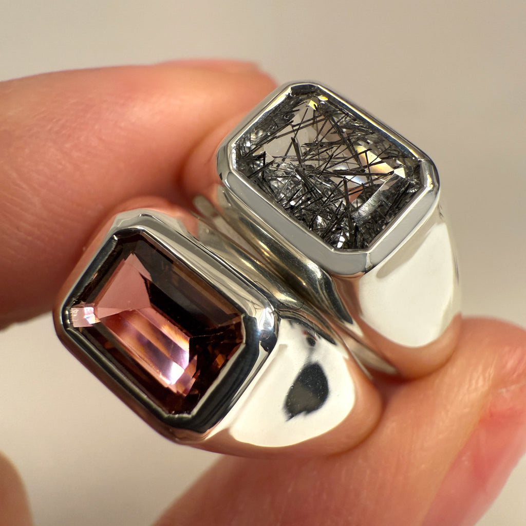 A photo of two silver signet rings with a black rutile quartz and a pink tourmaline  in between two fingers 