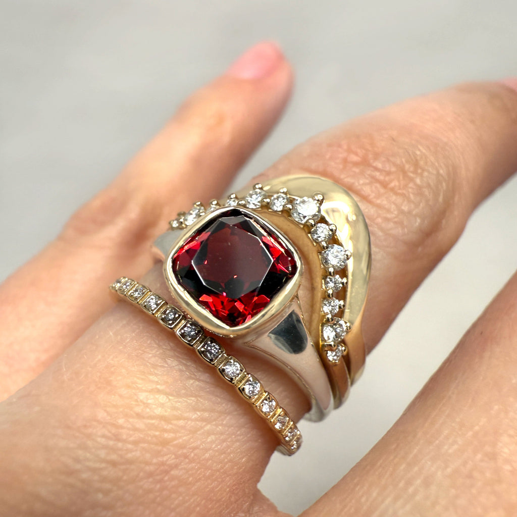 A finger wearing a red garnet gemstone ring set in silver and stacked with three other white diamond and yellow gold stacking bands 