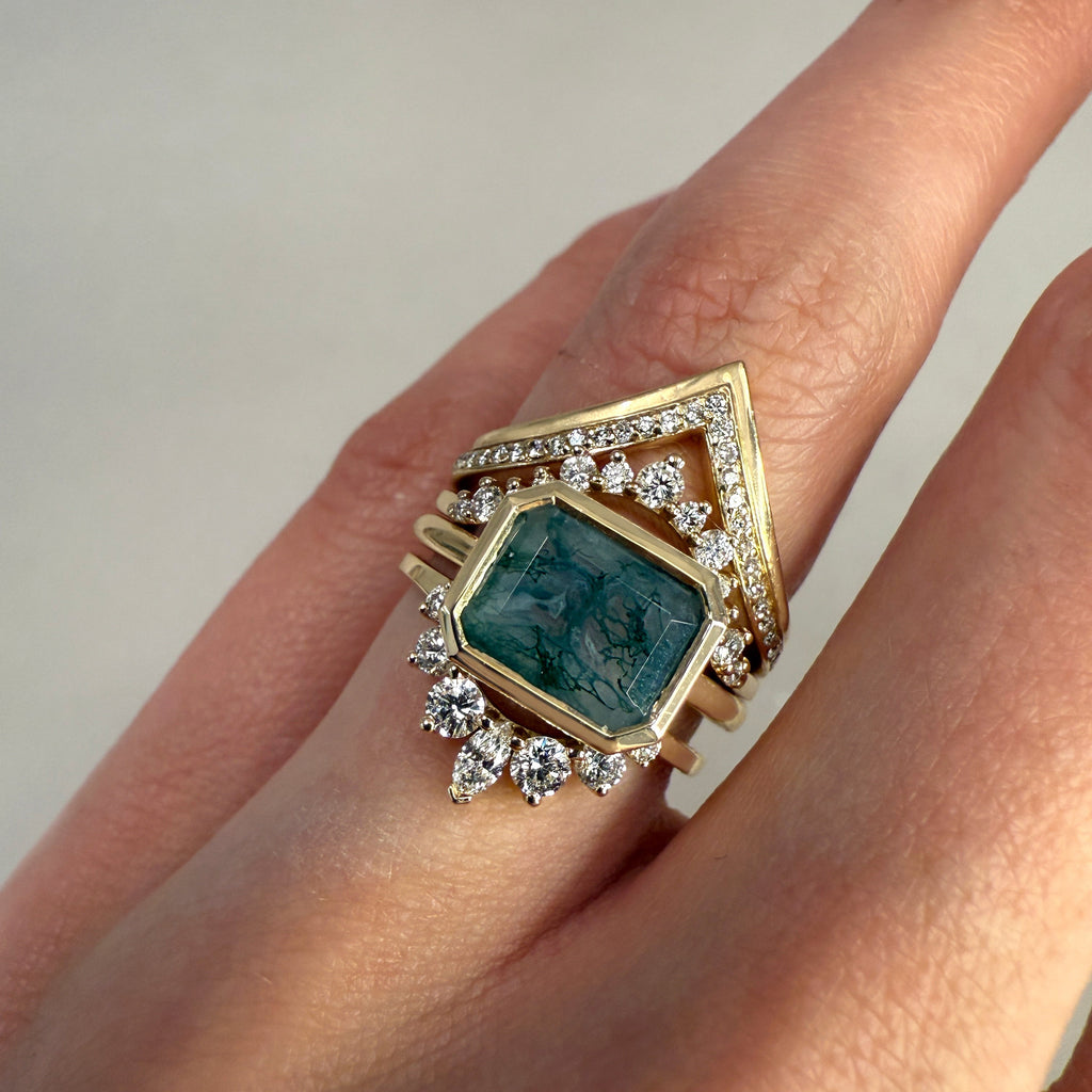 A ring wearing a green bezeled gemstone ring with white diamond stacking bands against on a grey background