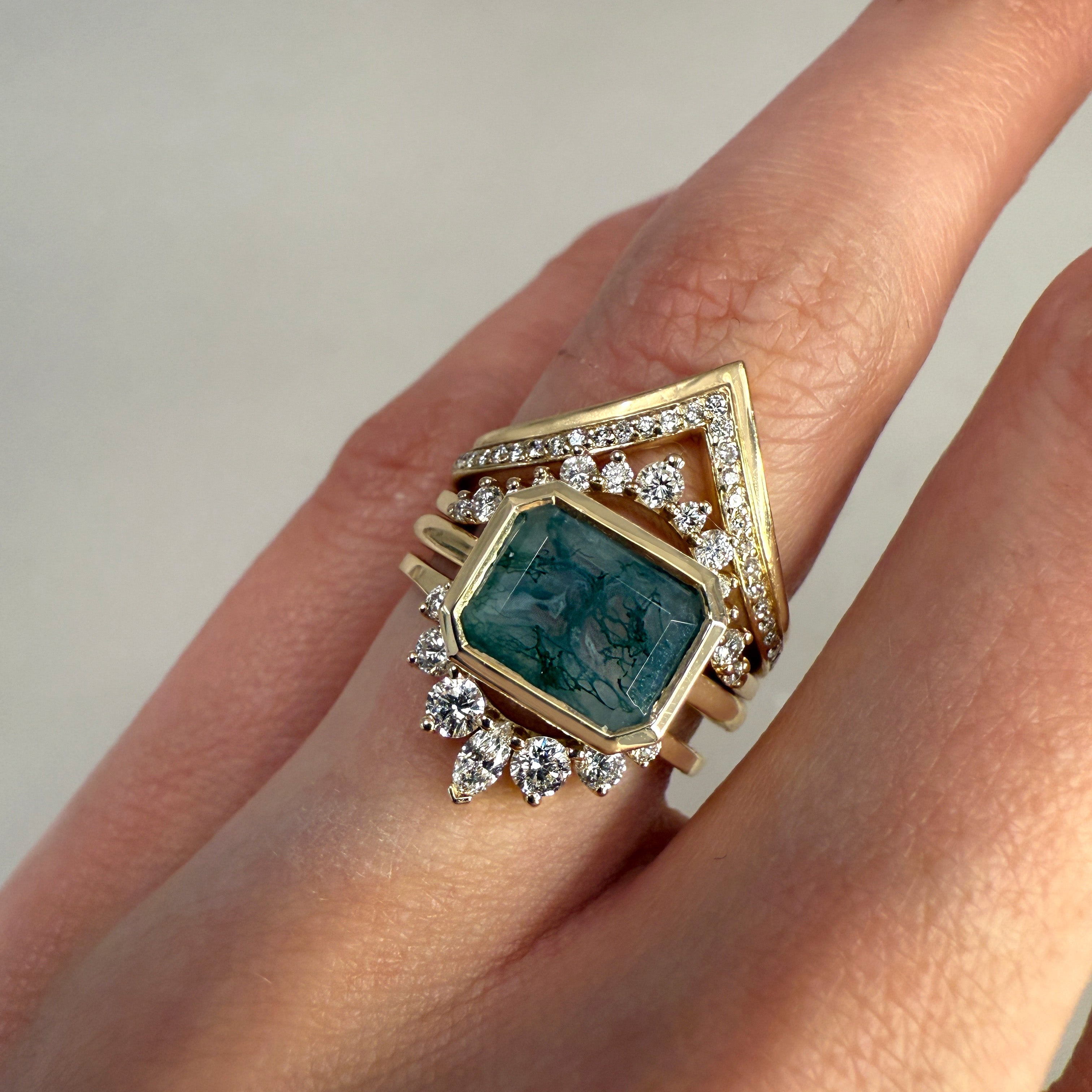 A ring wearing a green bezeled gemstone ring with white diamond stacking bands against on a grey background