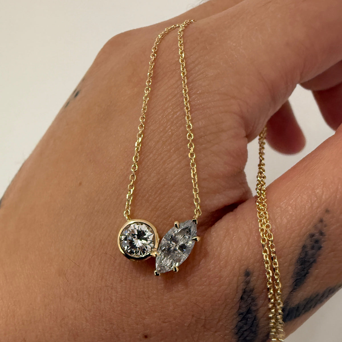 A close-up of a hand holding a delicate gold chain necklace with two pendants: a round bezel-set clear stone and an oval clear stone, against a plain light background.